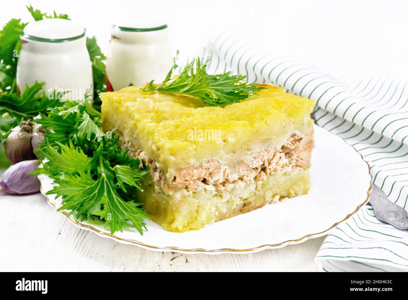 Mashed potato casserole with salmon fillet and lettuce in a plate, kitchen towel, garlic on the background of a light wooden board Stock Photo