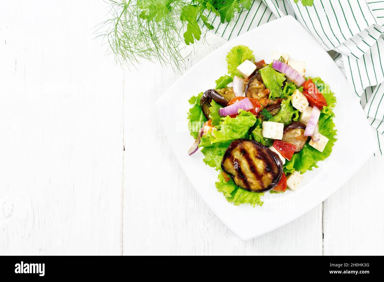 Salad of fried eggplant, fresh tomato with red onion and salted feta cheese, seasoned with vegetable oil and soy sauce on lettuce in plate, napkin, fo Stock Photo
