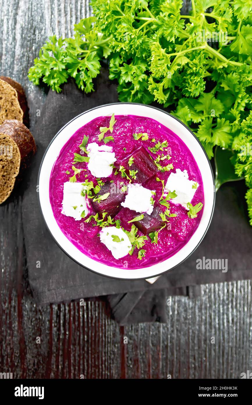 Soup puree of beetroot, potato and cream with salted cream cheese in a bowl on napkin, bread and parsley on dark wooden board background from above Stock Photo