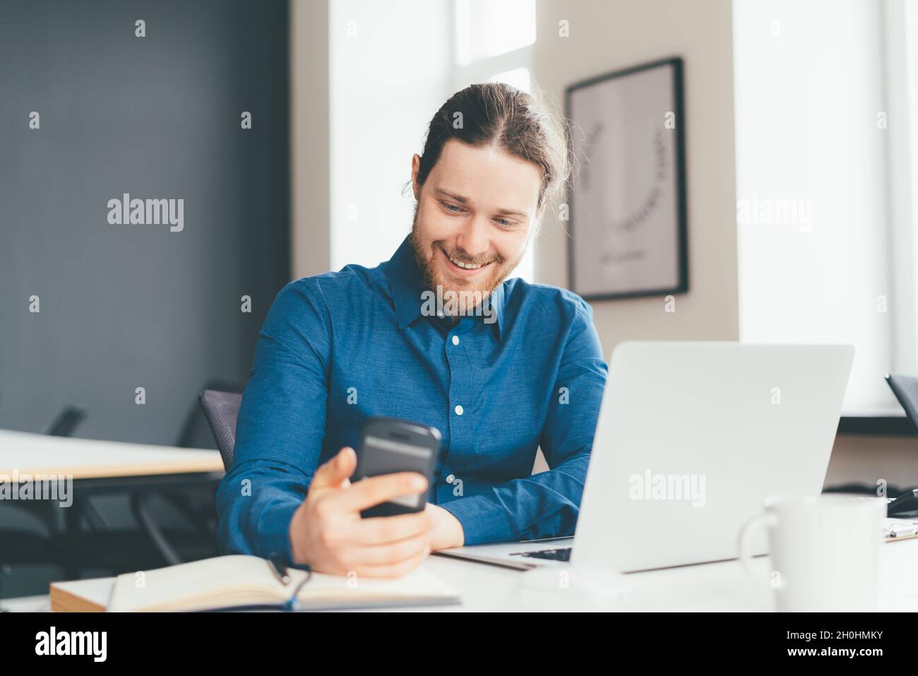 Smiling young male office worker using smartphone while working on laptop in modern office centre Stock Photo