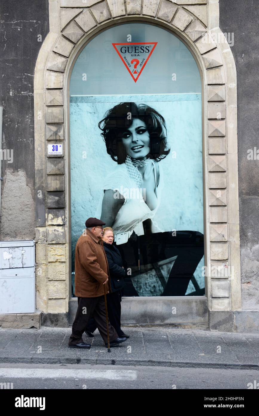 An elderly Sicilian couple walking under a large advertisement of Guess  clothing shop at the Palazzo Tezzano in Catania, Italy Stock Photo - Alamy