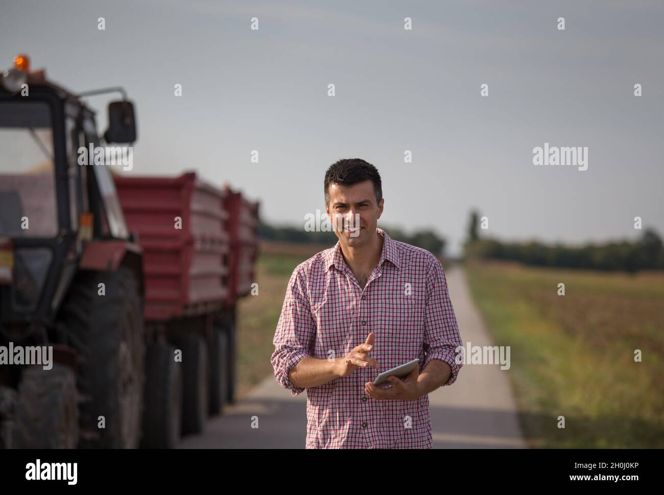 Handsome farmer with tablet standing in front of tractor with trailers ...