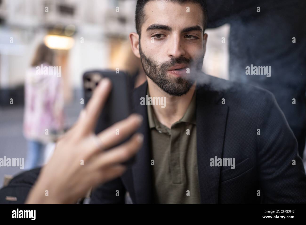 Sorry man blowing cigarette smoke while watching content on his girlfriend cellphone display. Woman discovers the betrayal through social networks and Stock Photo