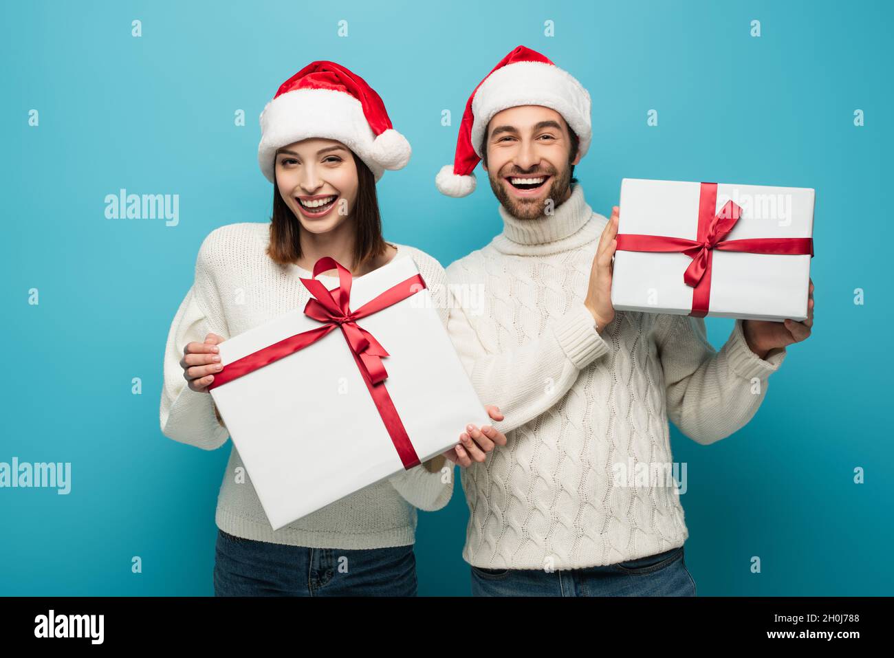 joyful couple in white sweaters and santa hats holding presents and looking at camera on blue Stock Photo