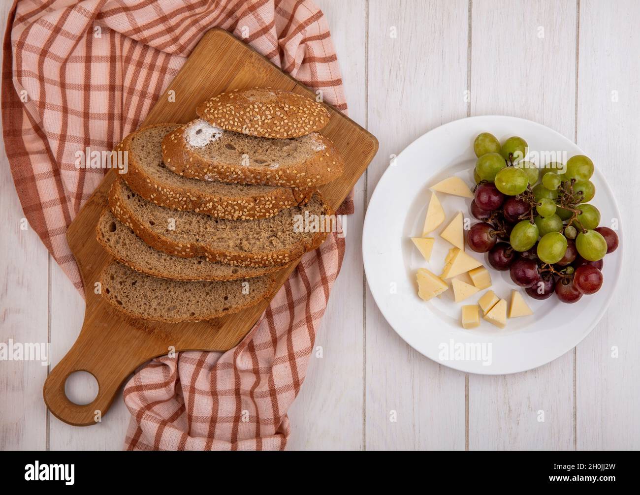 top view of brown sliced seeded cob on cutting board on plaid cloth and plate of cheese and grape on wooden background Stock Photo