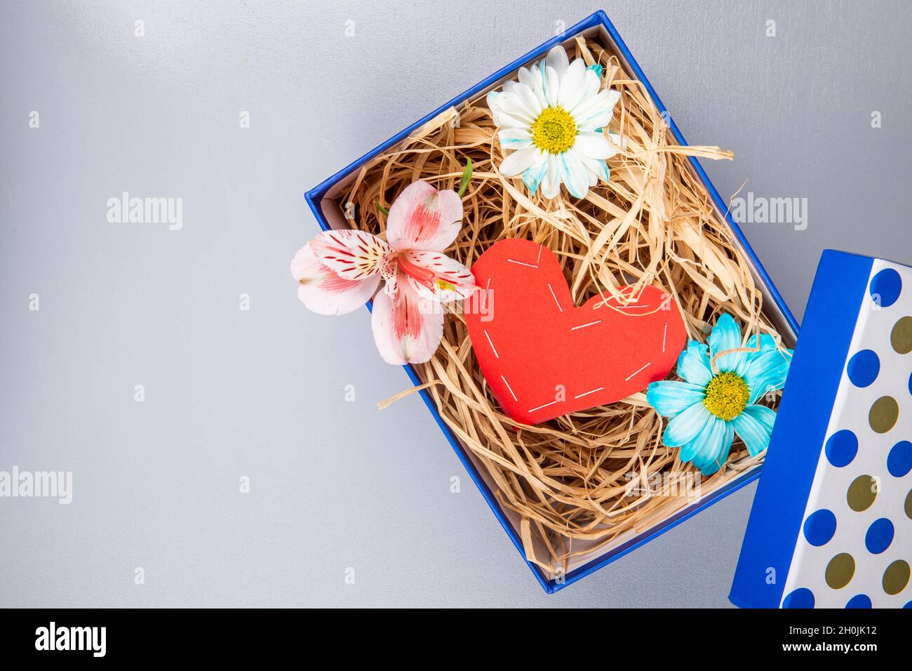 top view of colorful daisy flowers and pink alstroemeria with a heart made from red color paper and with straw in a blue present box on white backgrou Stock Photo