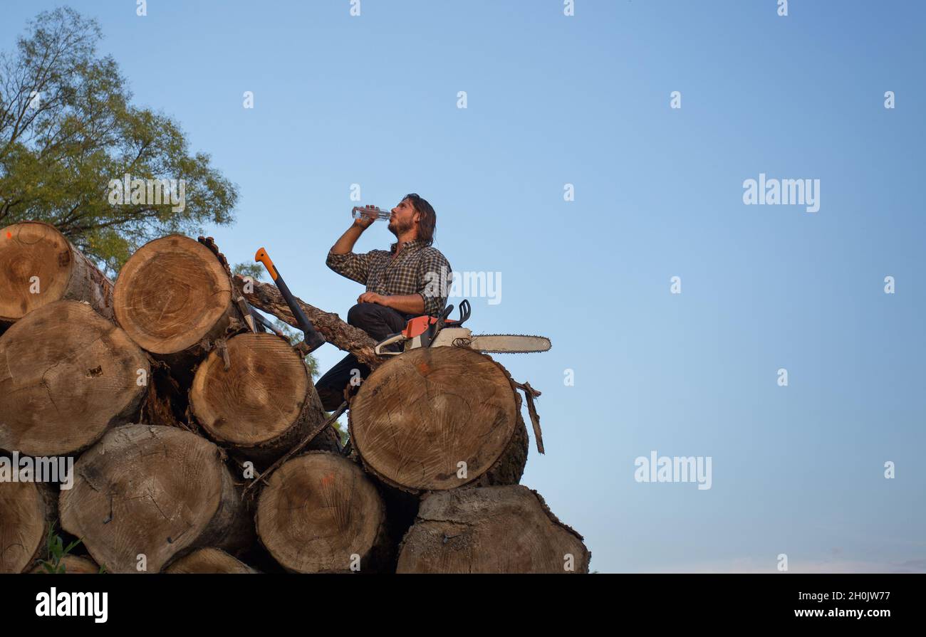 Young strong lumberjack sitting on pile of trunks with chainsaw and ax beside and drinking water from bottle Stock Photo