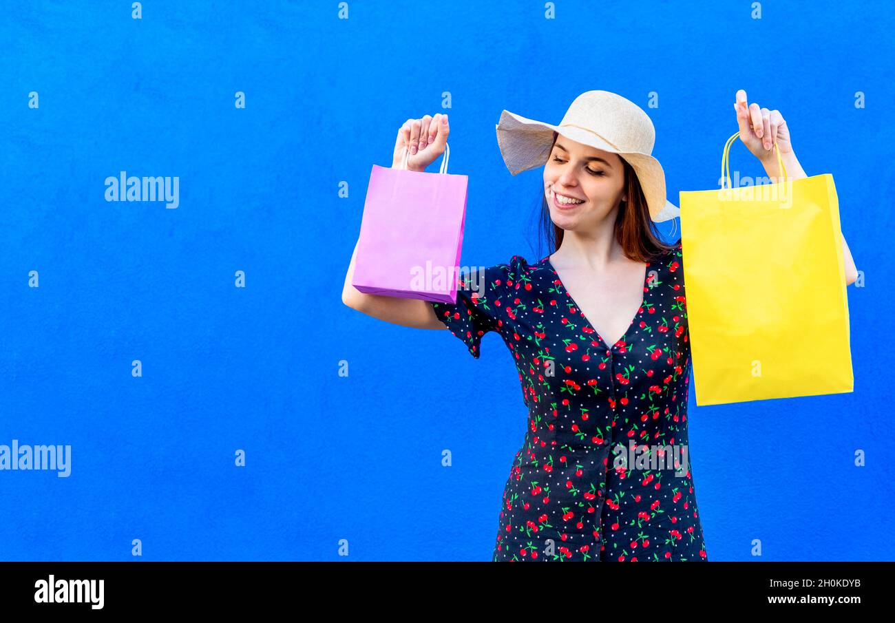 young happy woman wearing summer clothes holding shopping bags isolated on a blue background wall. girl shop gifts on black friday celebrating sales. Stock Photo
