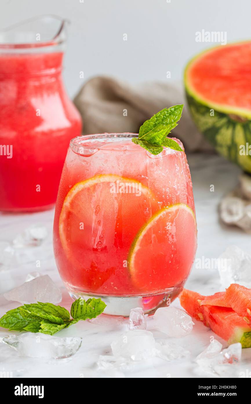 Watermelon mojito fresh red drink. With watermelon, lemon, lime, mint leaves and ice. The drink is on a white table with a half watermelon in the back Stock Photo