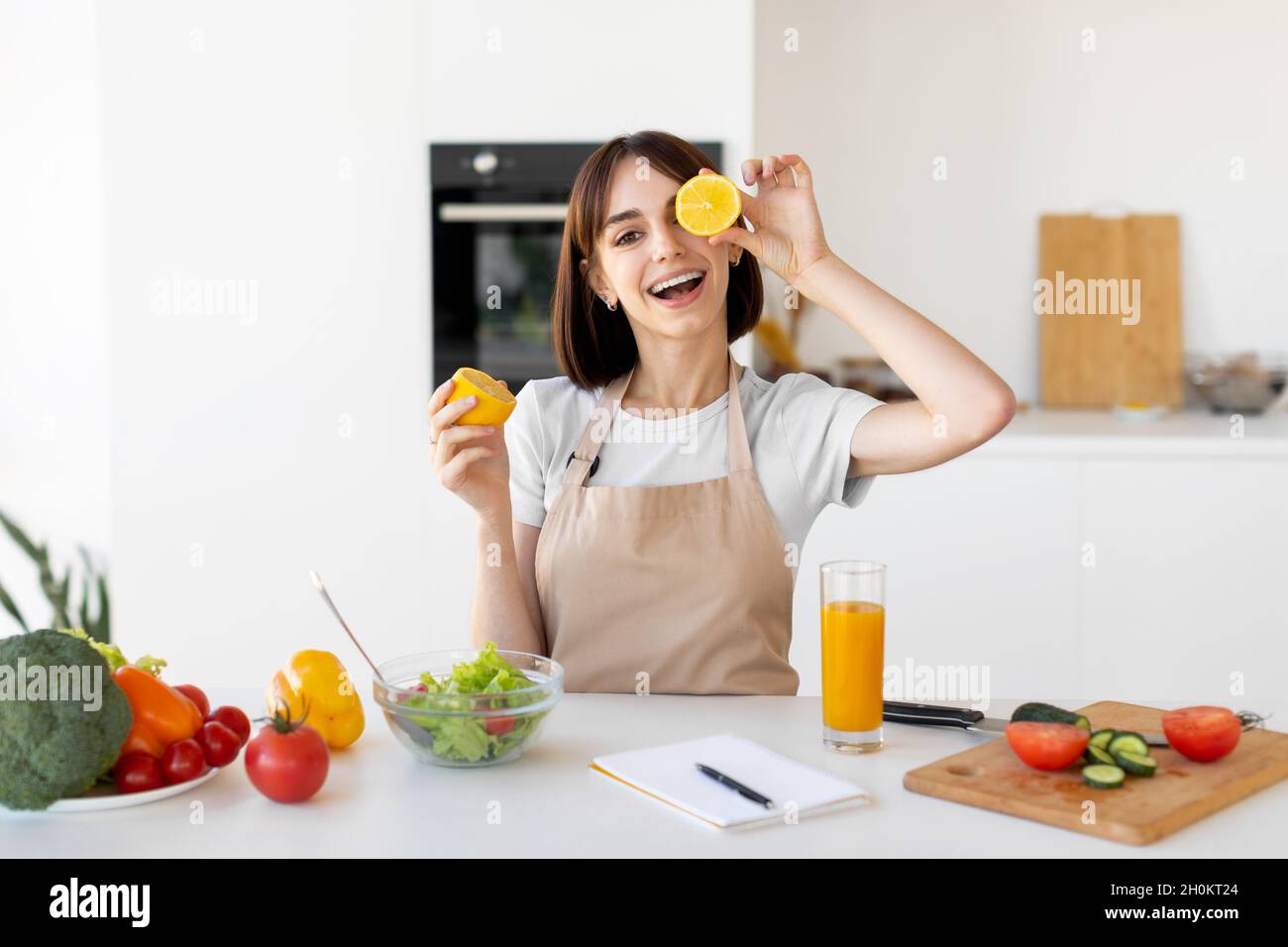 Playful young woman having fun and covering her eyes with lemon slices, fooling around in kitchen interior Stock Photo