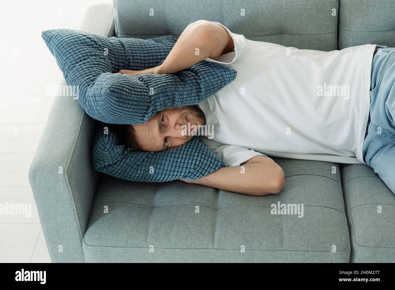Annoyed man cover ears with pillows to avoid noise from neighbor while napping on couch try to