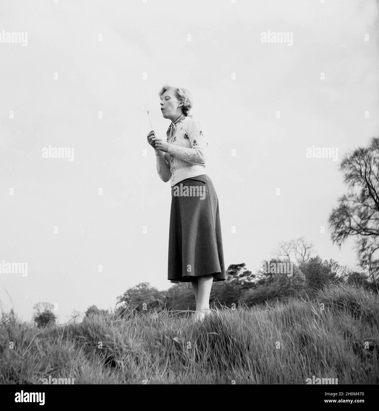 1950s, historical, a lady standing outside on a hillside blowing a dandelion flower and perhaps making a wish, England, UK. For centuries ancient folklore superstitions have been attached to the flower and many believe that blowing the seeds can grant your wishes but also carry your thoughts and dreams to those you love as they travel through the air. Stock Photo