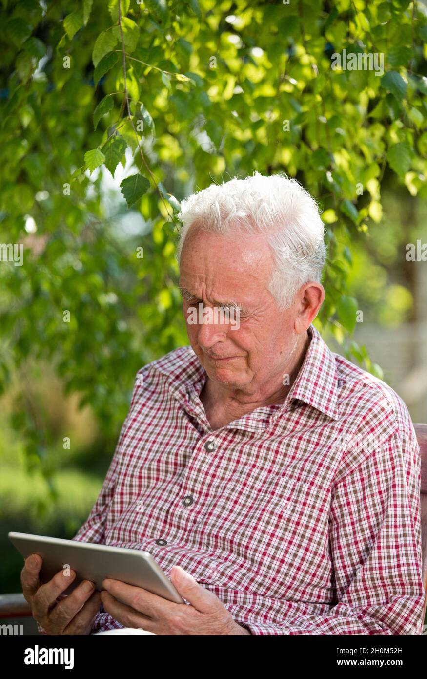 Sad senior man sitting on bench in garden, looking at tablet and crying Stock Photo