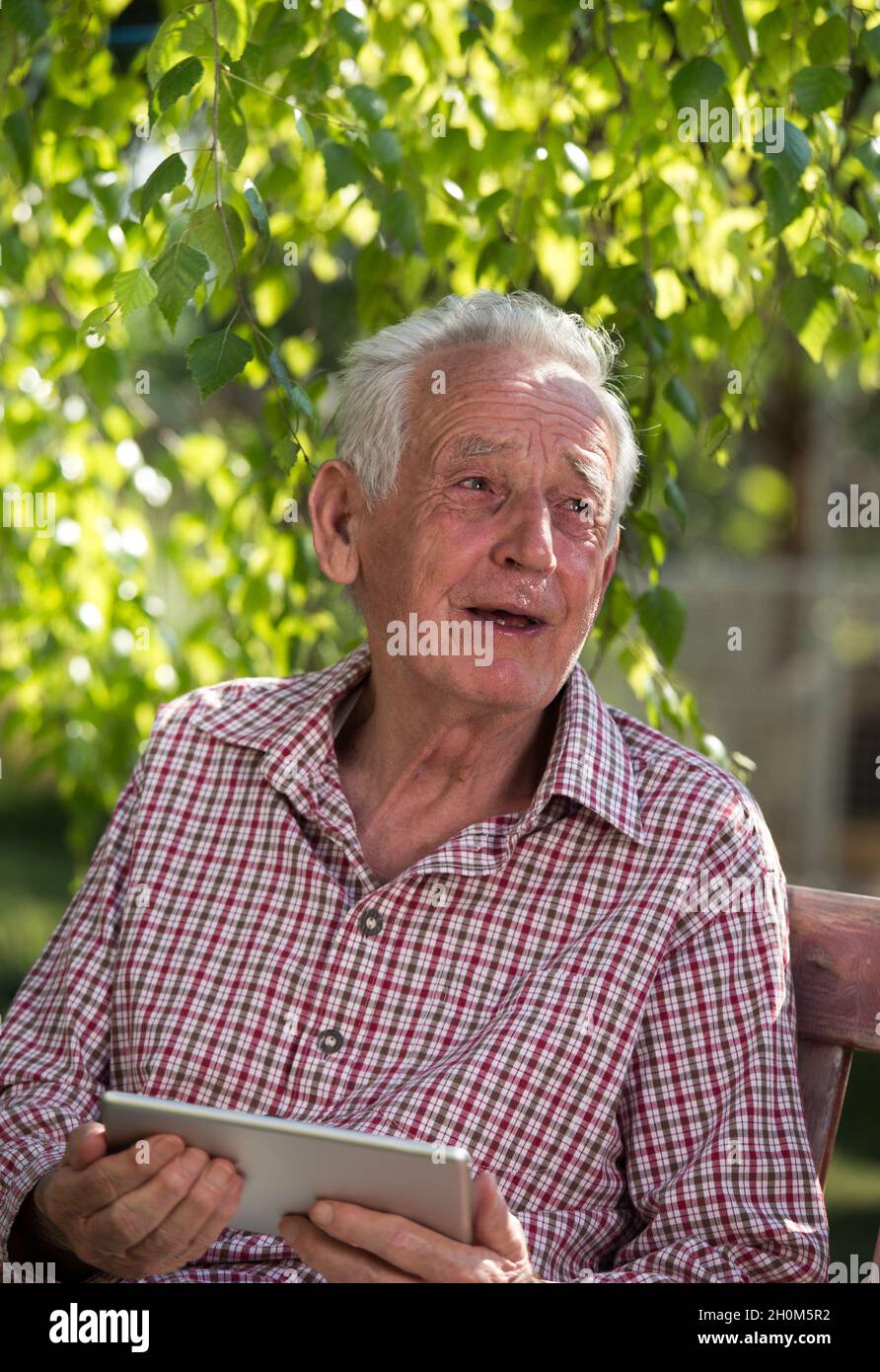 Sad senior man sitting on bench in garden, looking at tablet and crying Stock Photo