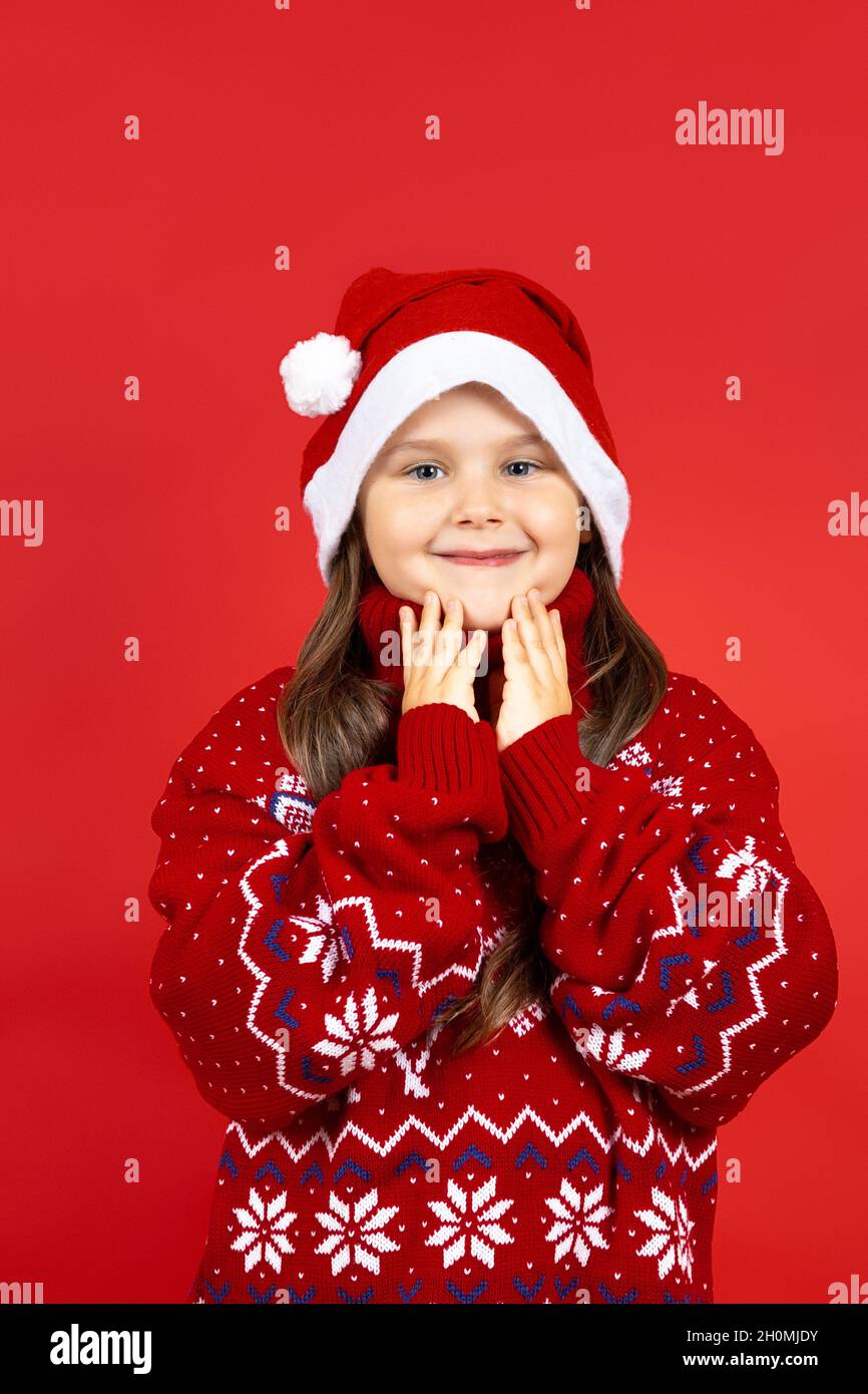 portrait of smiling, charming girl in red knitted Christmas sweater with reindeer touching her face with palms, isolated on a red background Stock Photo