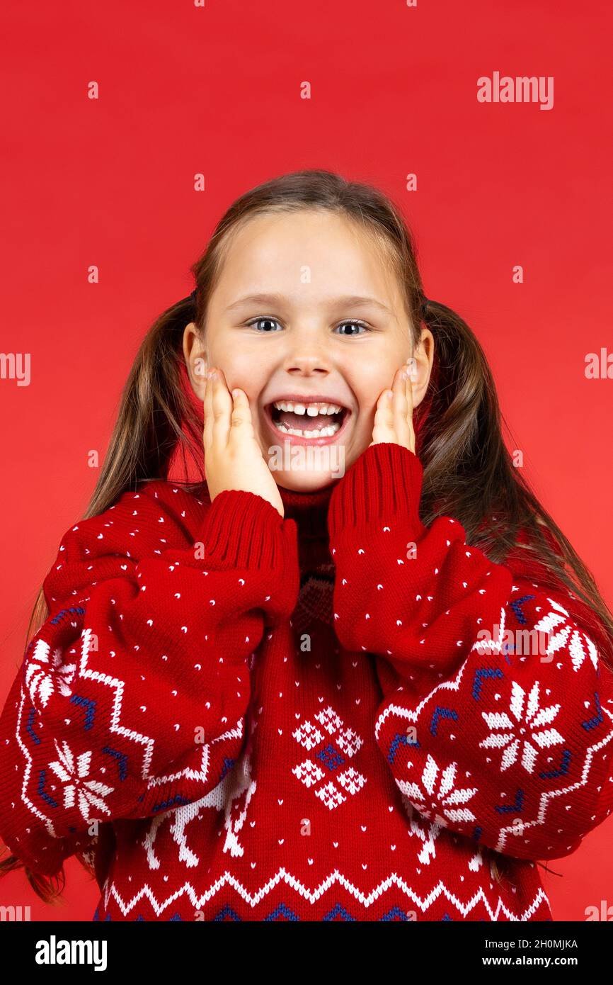 portrait of laughing, surprised girl in red Christmas sweater with reindeer touching face with palms, isolated on red background Stock Photo