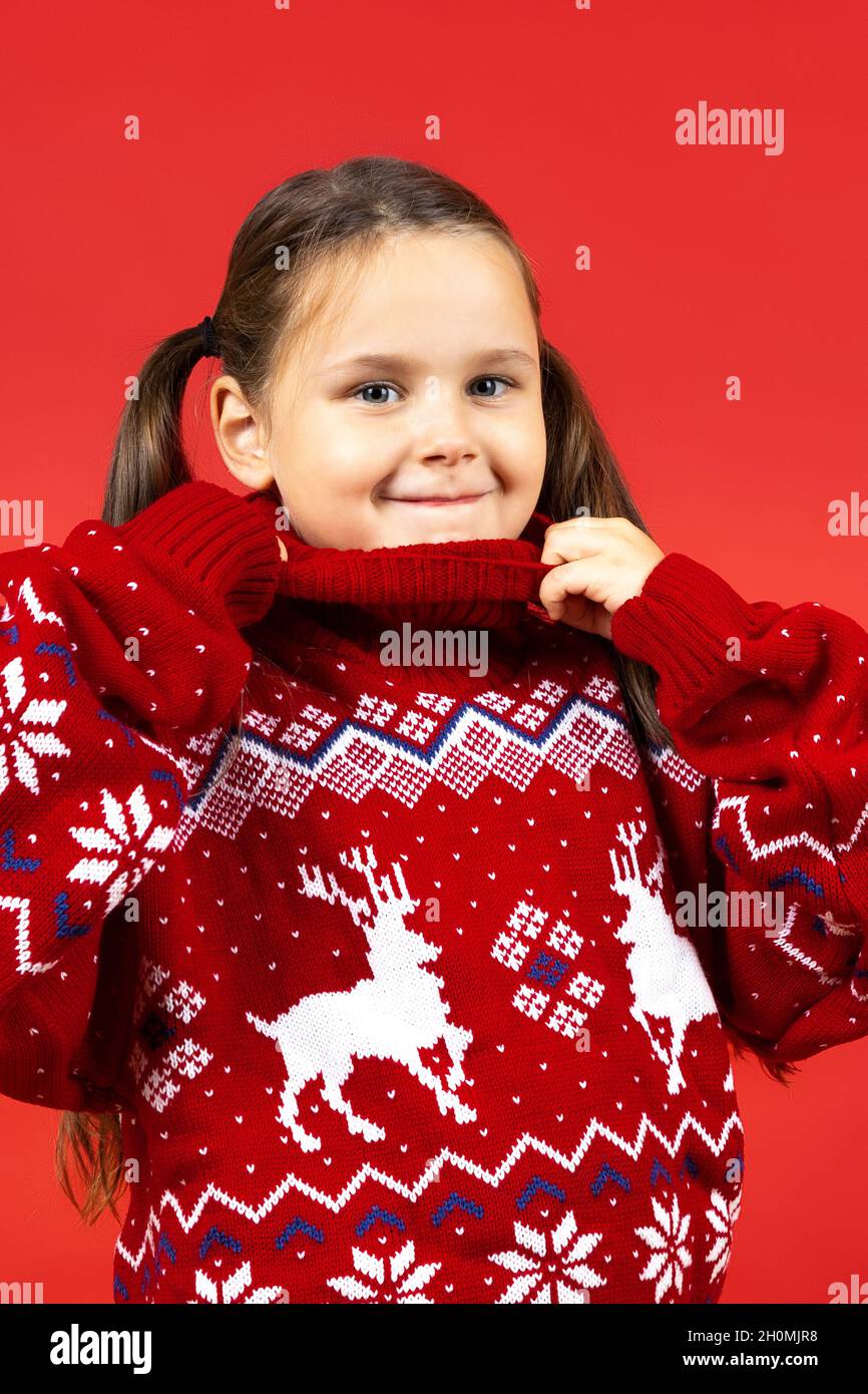 close-up portrait of cute girl in red Christmas sweater with reindeer holding collar with hands, isolated on red background Stock Photo