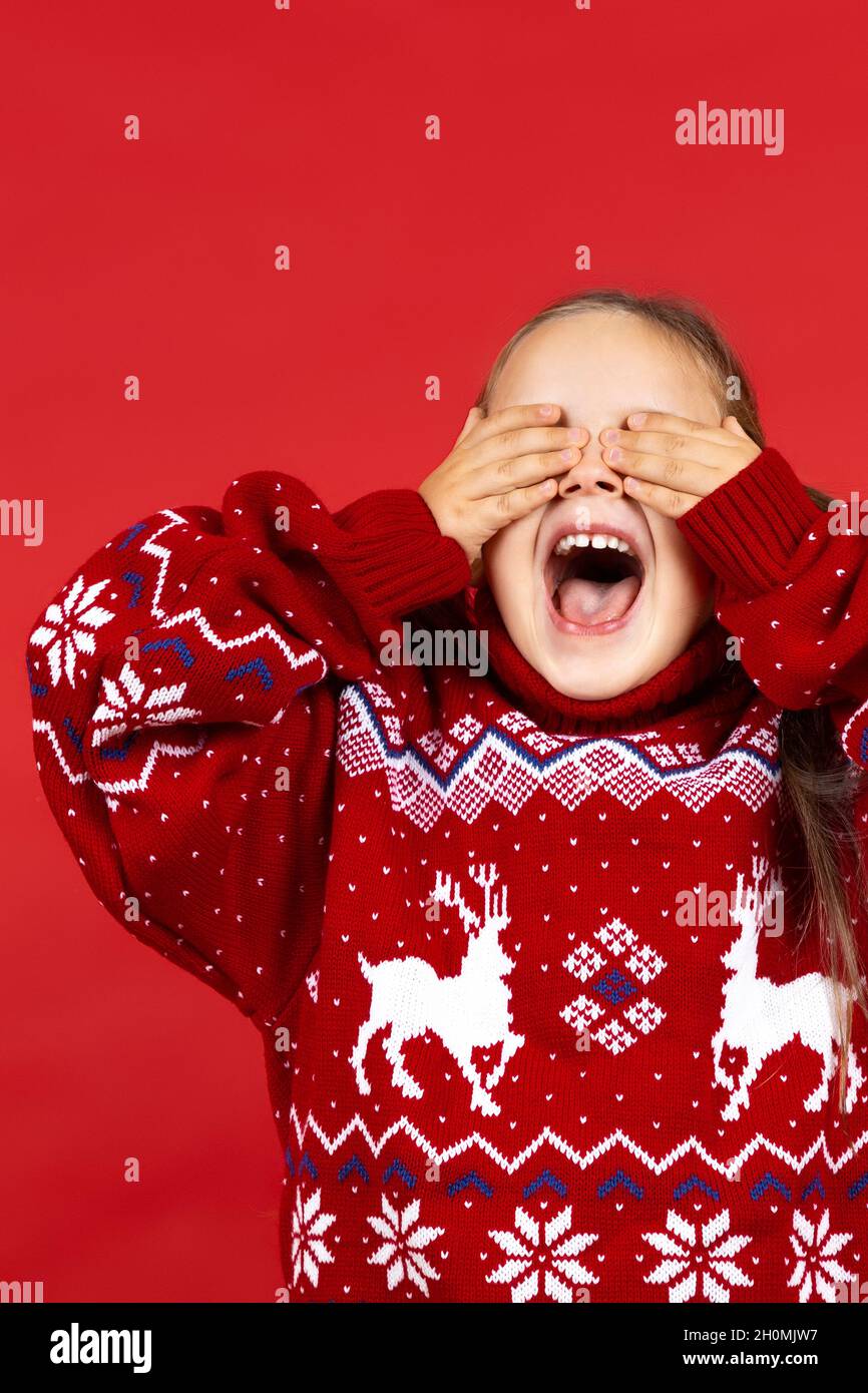 portrait of laughing, positive girl in red Christmas sweater with reindeer covering eyes with hands in anticipation of surprise, isolated on red Stock Photo