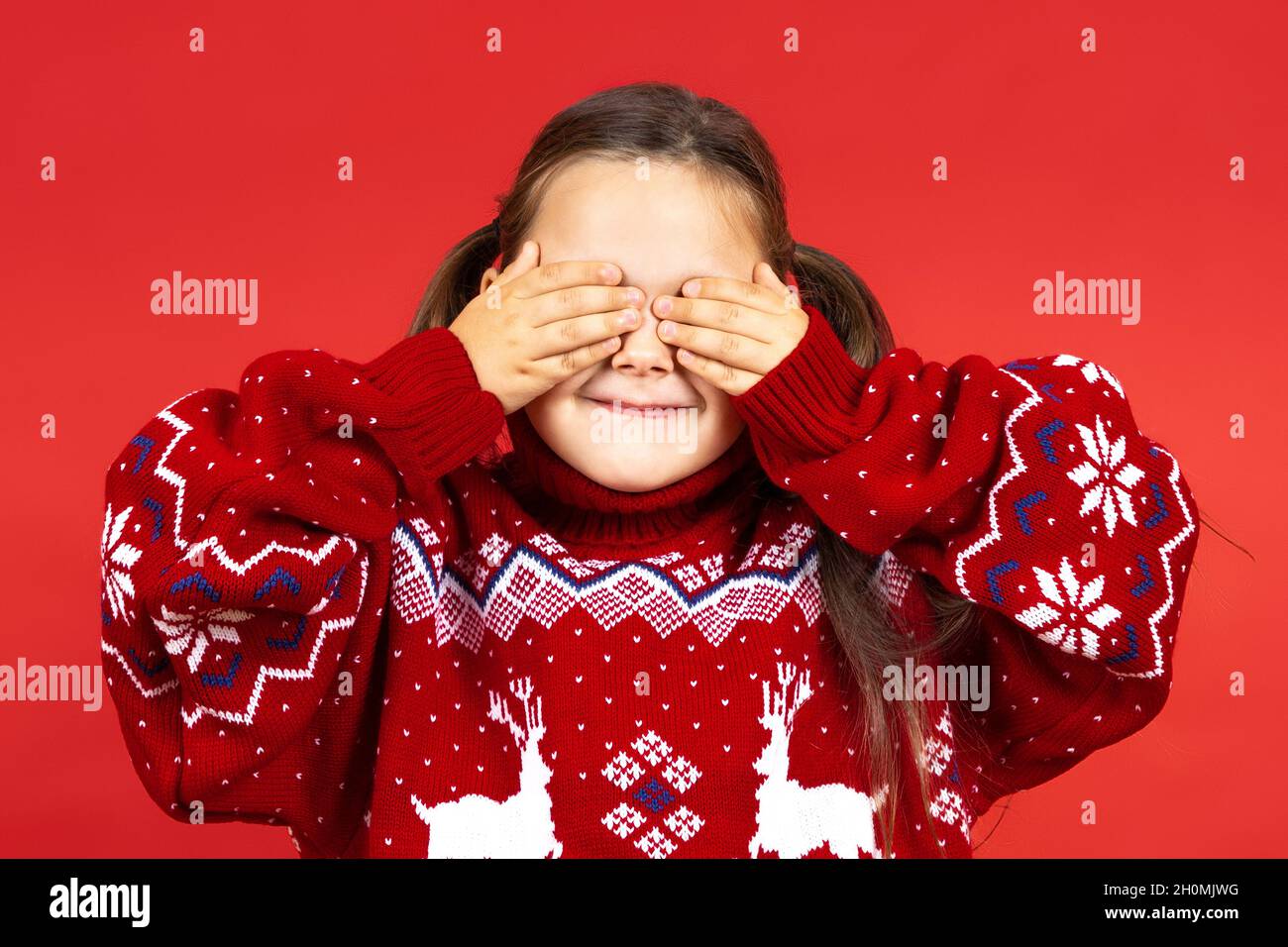 close-up portrait of smiling, charming girl in red Christmas sweater with reindeer covering her eyes with hands and waiting for gift, isolated on red Stock Photo
