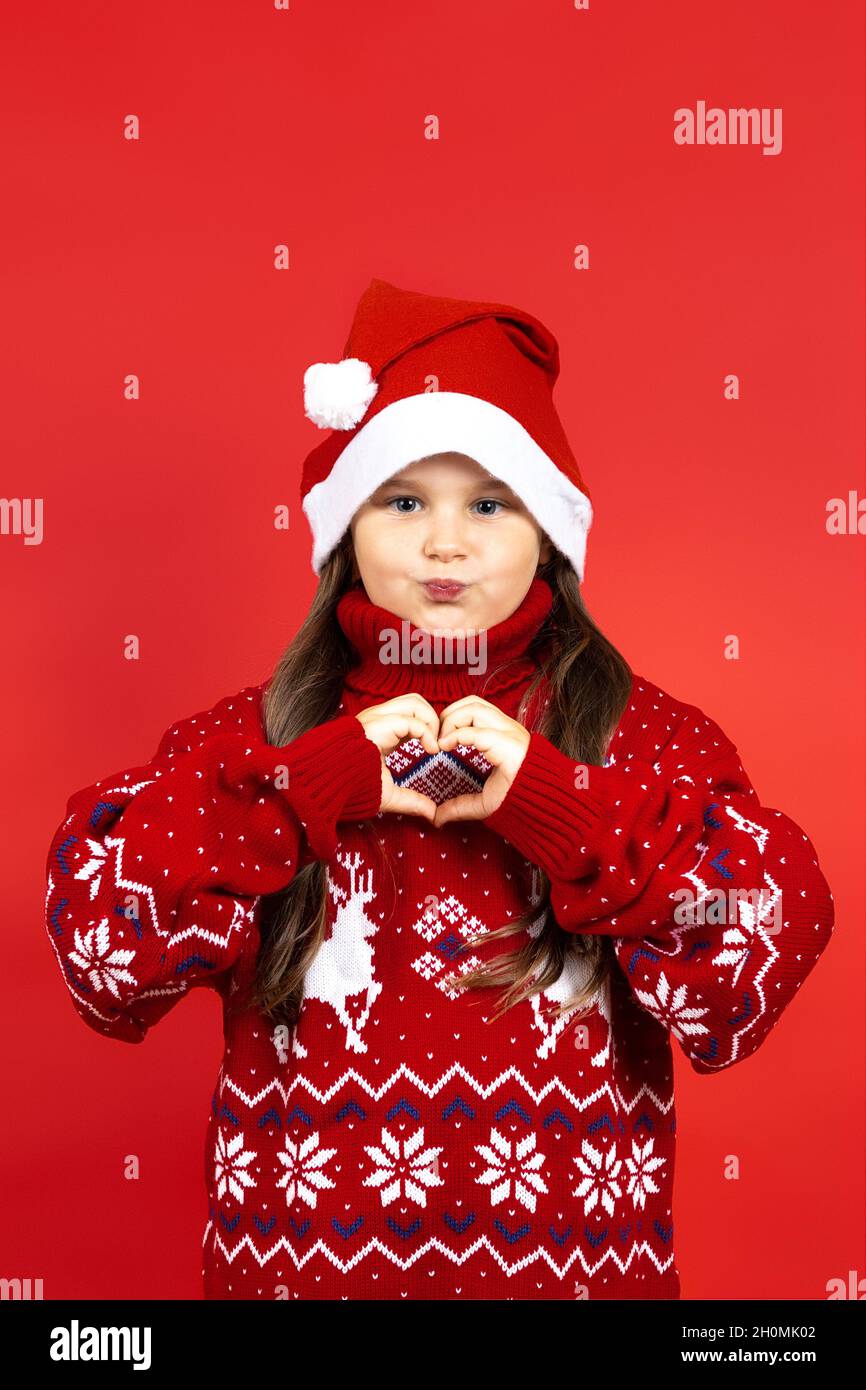 portrait of mischievous, playful girl in red knitted Christmas sweater with reindeer and red hat showing heart sign with fingers, isolated on red Stock Photo