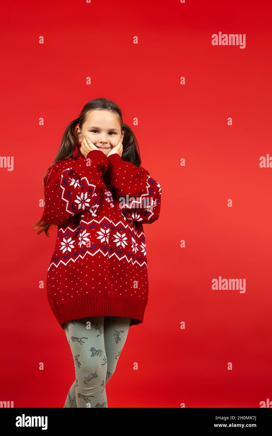 full-length portrait of cute girl in red knitted Christmas sweater with reindeer touching face with hands, isolated on red background Stock Photo