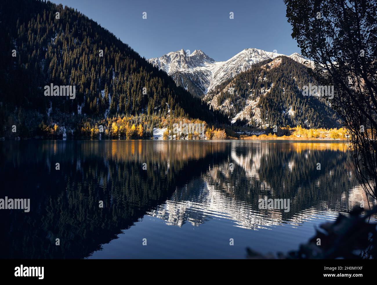 Mountain landscape of autumn forest with yellow trees and snowy white peak with reflection on Lake Issyk in Kazakhstan Stock Photo