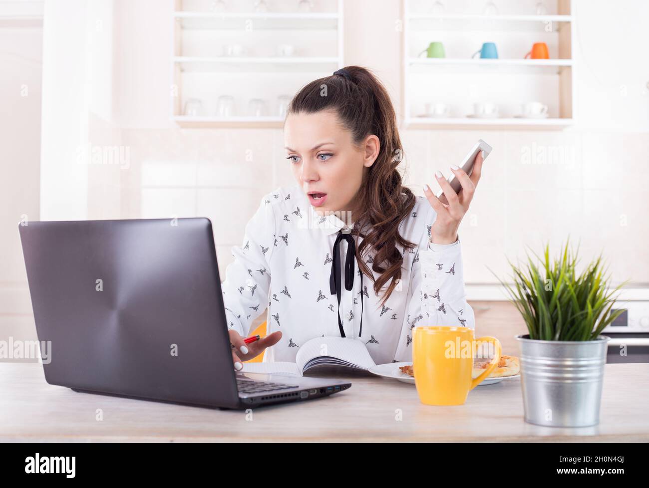 Shocked young woman holding mobile phone and looking at laptop. Busy girl working from home Stock Photo