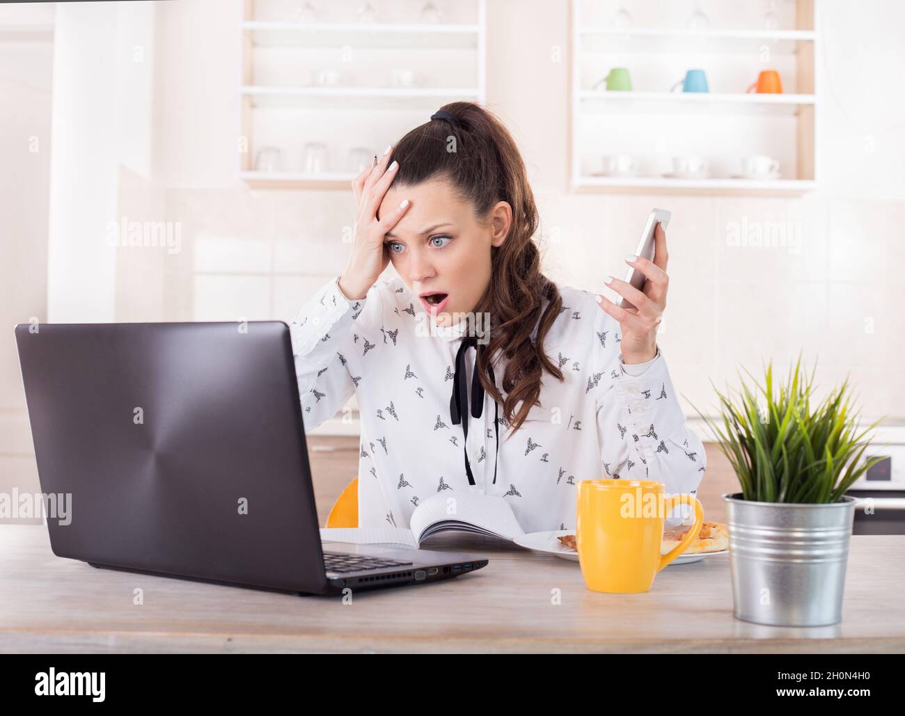 Shocked young woman holding mobile phone and looking at laptop. Busy girl working from home Stock Photo