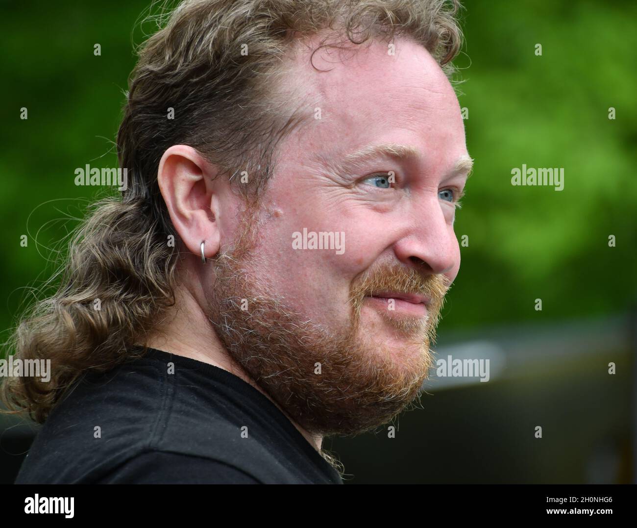 White male with mullet hair and smile in front of green trees ...