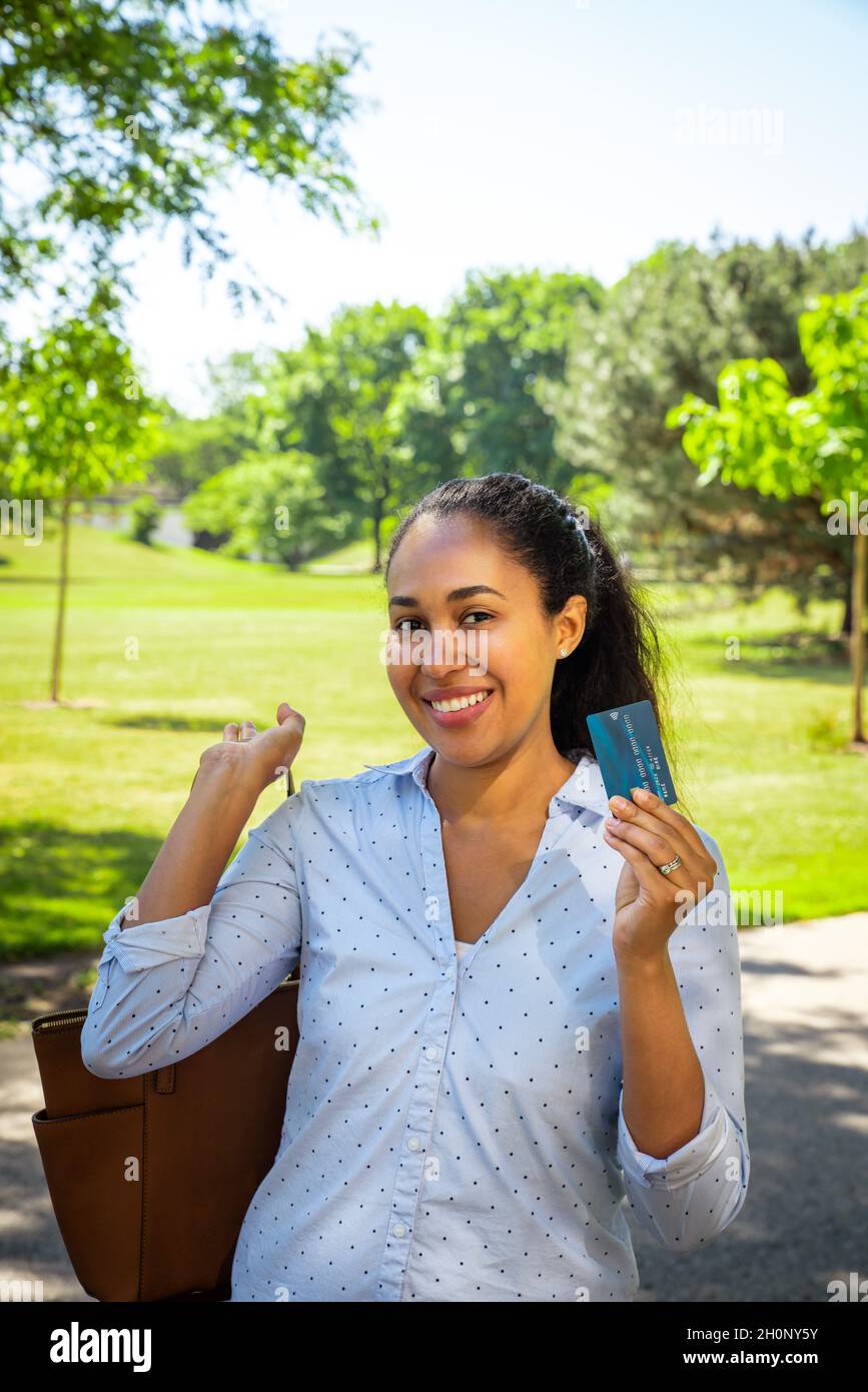 A smiling mixed race African American woman in a blue collared shirt holds up a blue plastic credit card with micro chip with her brown purse slung ov Stock Photo