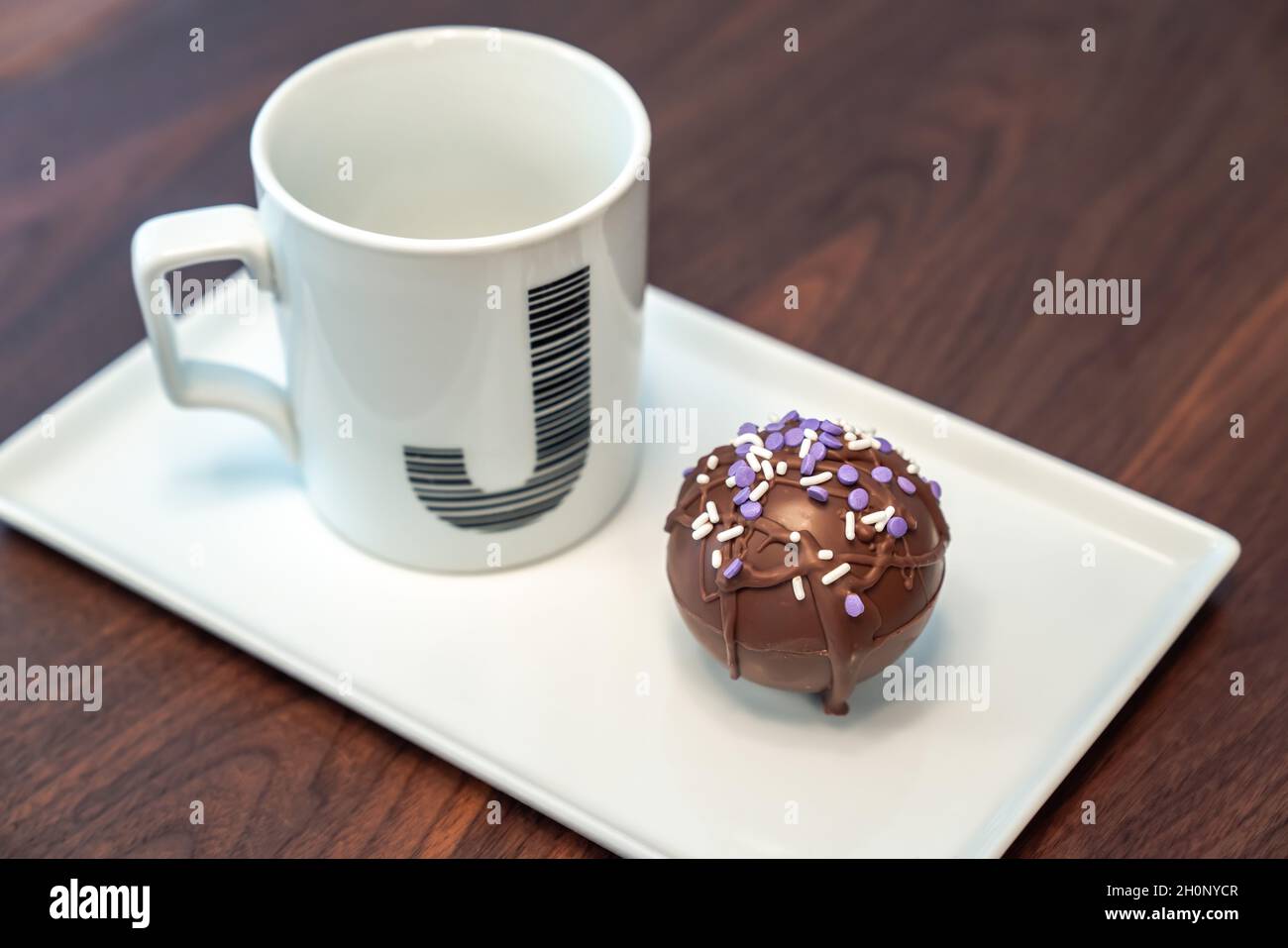 Close up food photograph of a hot cocoa bomb covered in drizzled brown chocolate and purple and white sprinkles set on a white serving plate next to a Stock Photo