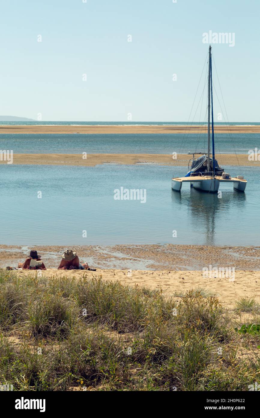 A couple reading books on beach in front of their anchored boat. Stock Photo