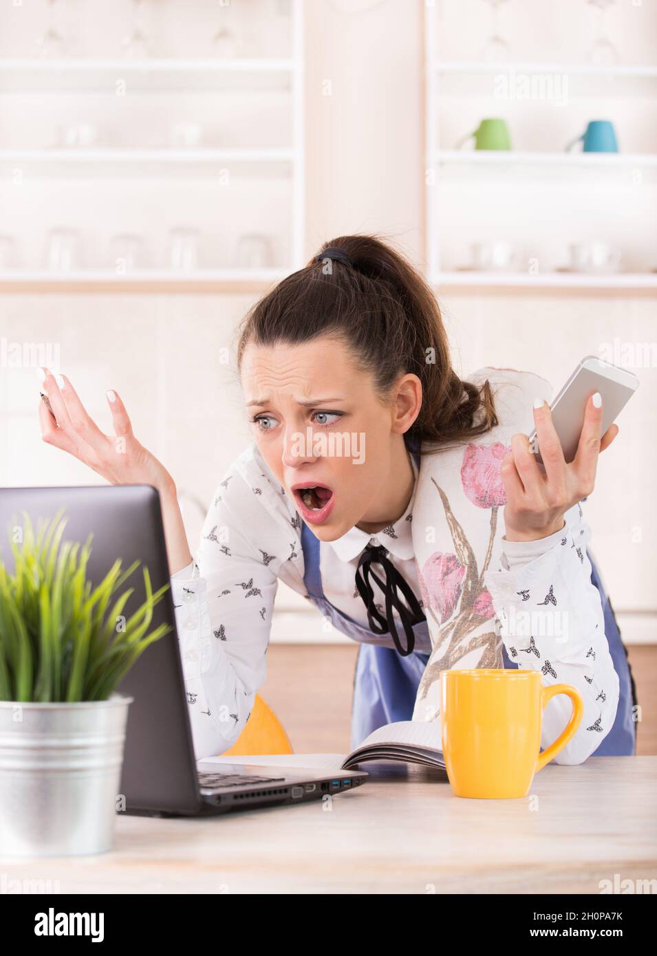 Shocked young woman holding mobile phone and looking at laptop. Busy girl with apron cooking and working from home Stock Photo