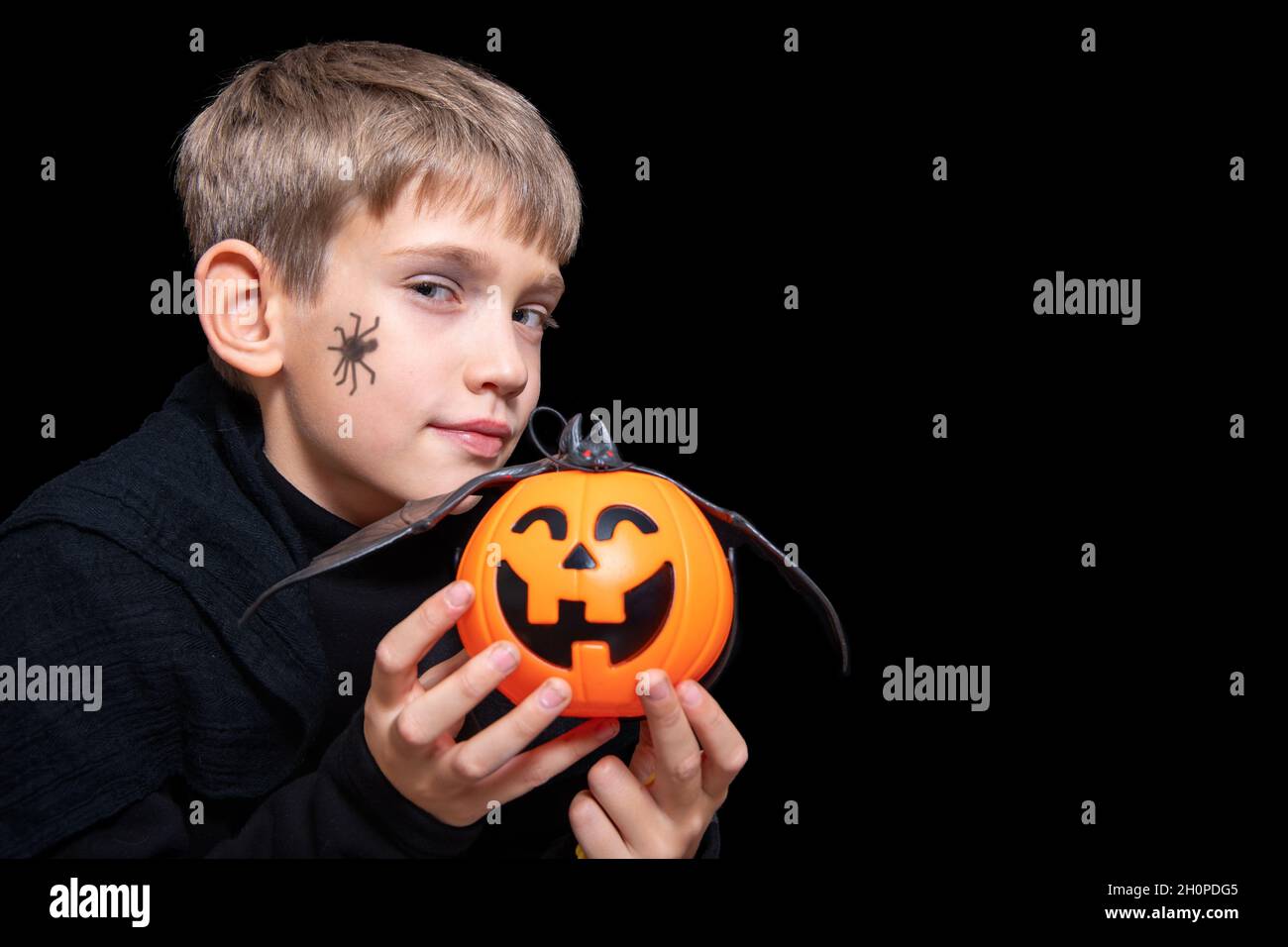 A child holding an orange pumpkin-shaped basket with a grinning face, Jack's lantern and a bat. A boy with a spider on his cheek is waiting for Hallow Stock Photo