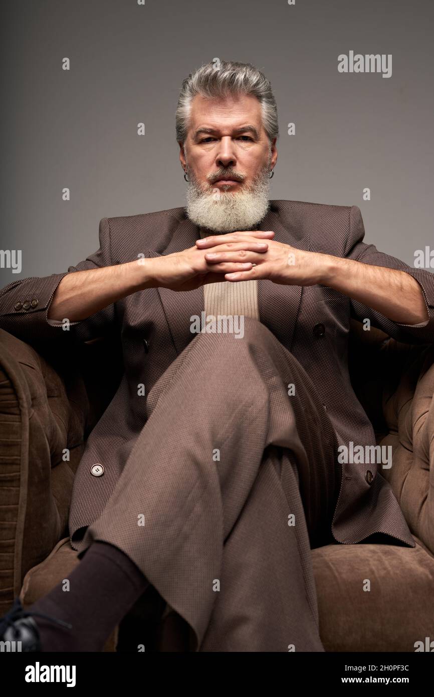 Portrait of serious mature businessman with beard wearing elegant suit looking at camera, sitting with hands crossed in armchair while posing isolated Stock Photo