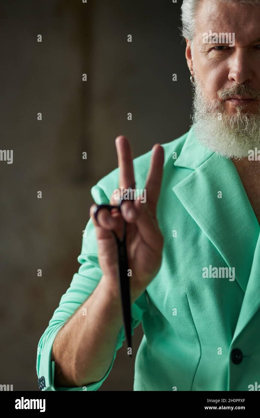 Close up shot of stylish middle aged man with beard wearing colorful outfit looking at camera, showing peace sign while holding sharp barber scissors Stock Photo