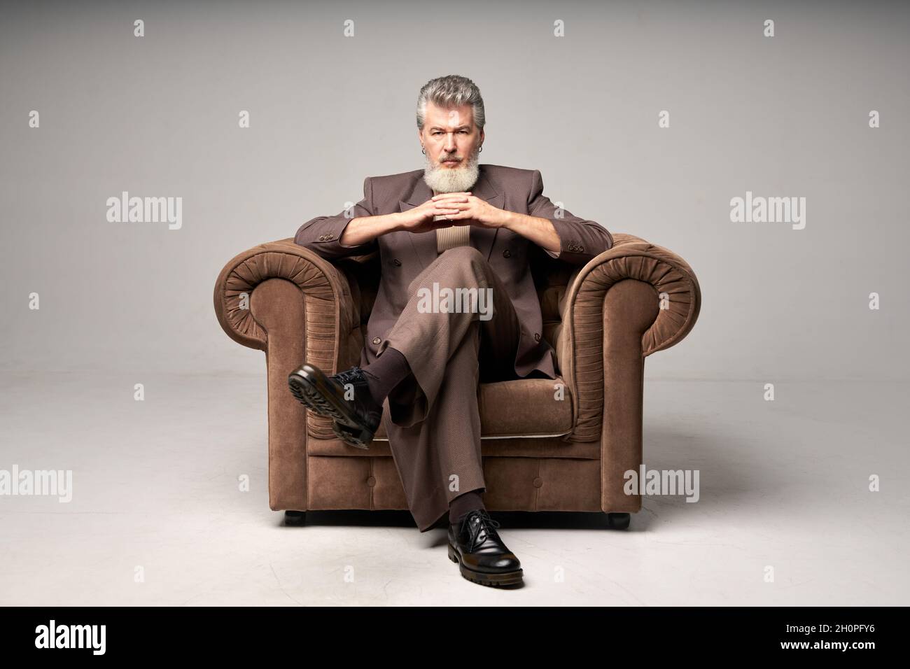 Full length shot of brutal mature businessman with beard wearing elegant suit looking at camera, sitting with hands crossed in armchair while posing Stock Photo