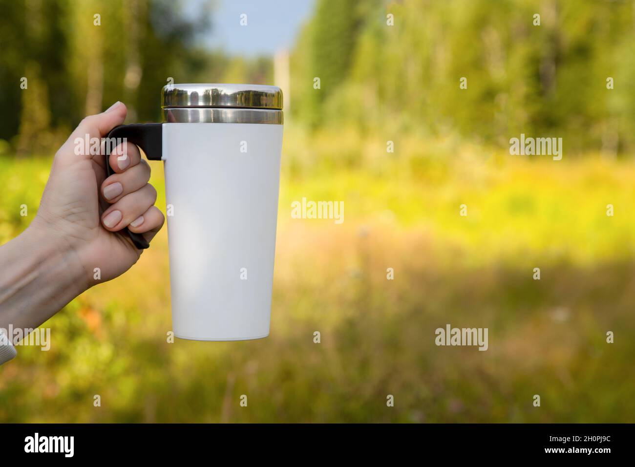 Mockup of a woman holding a white travel mug by a forest. Empty mug mock up for design promotion. Stock Photo