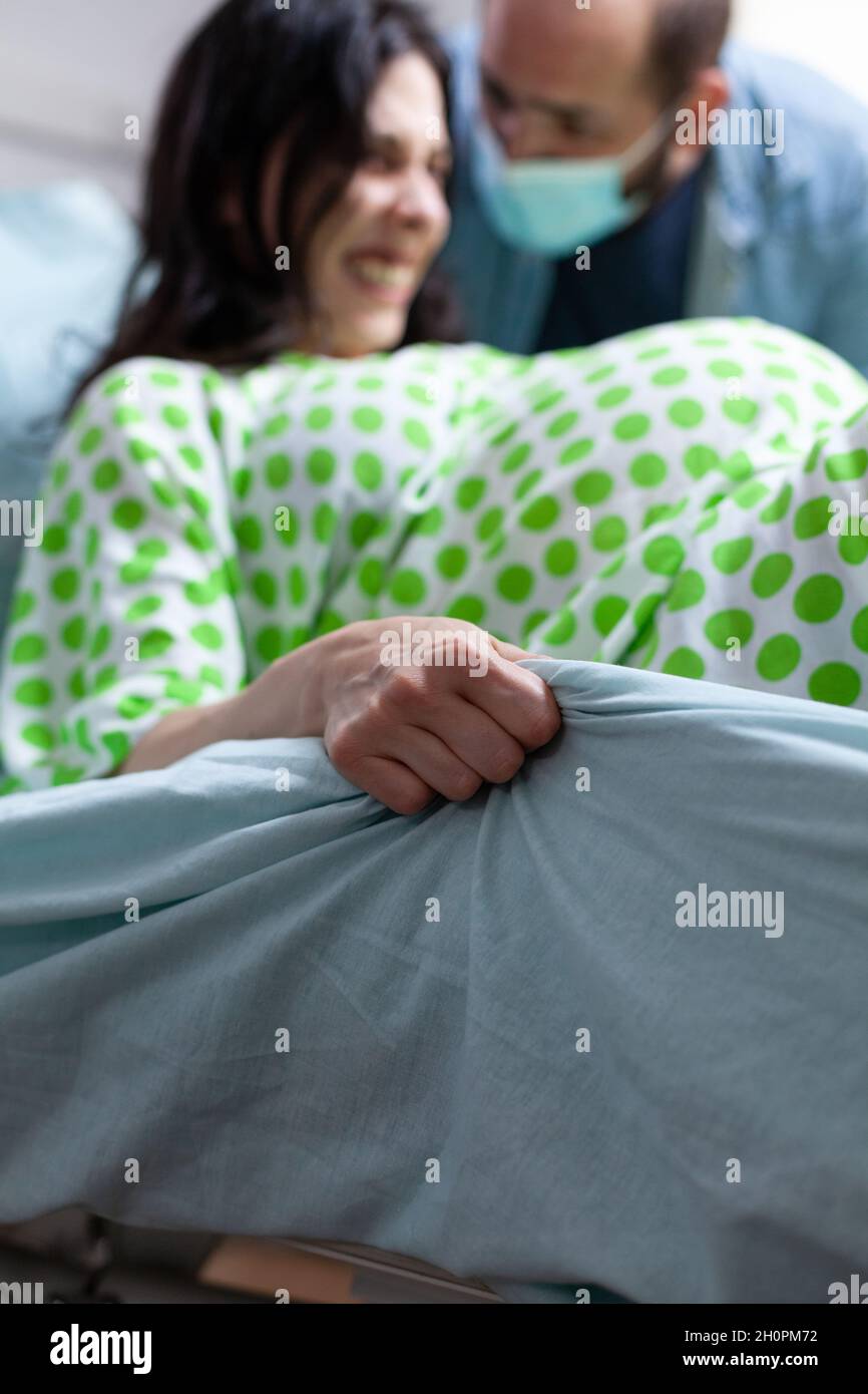 Close up of hand grabbing bedsheet from painful labor contractions at maternity. Young woman giving birth to child while father of baby supporting wife in hospital ward background Stock Photo