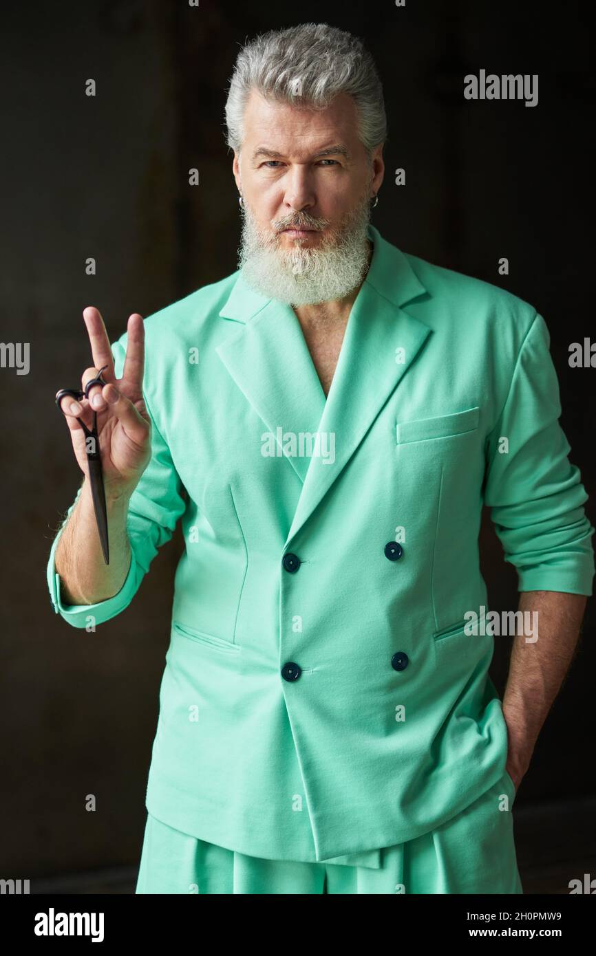 Cool gray haired middle aged man with beard wearing colorful outfit looking at camera, showing peace sign while holding sharp barber scissors, posing Stock Photo