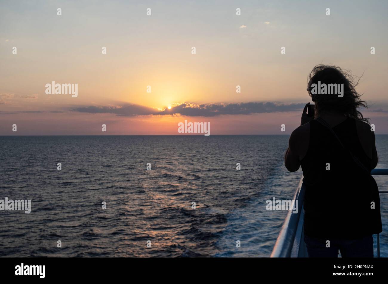 Sunset over the Mediterranean sea viewed from the ferry connecting Toulon and Corsica. Woman on the deck viewed from behind taking a picture of the su Stock Photo