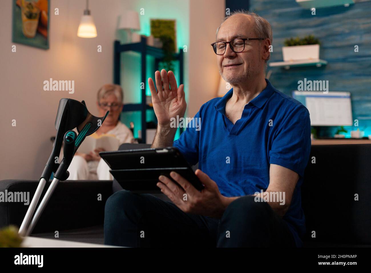 Elderly person waving at tablet with video call conference on sofa with crutches. Old man using online remote communication while senior woman sitting in wheelchair reading Stock Photo