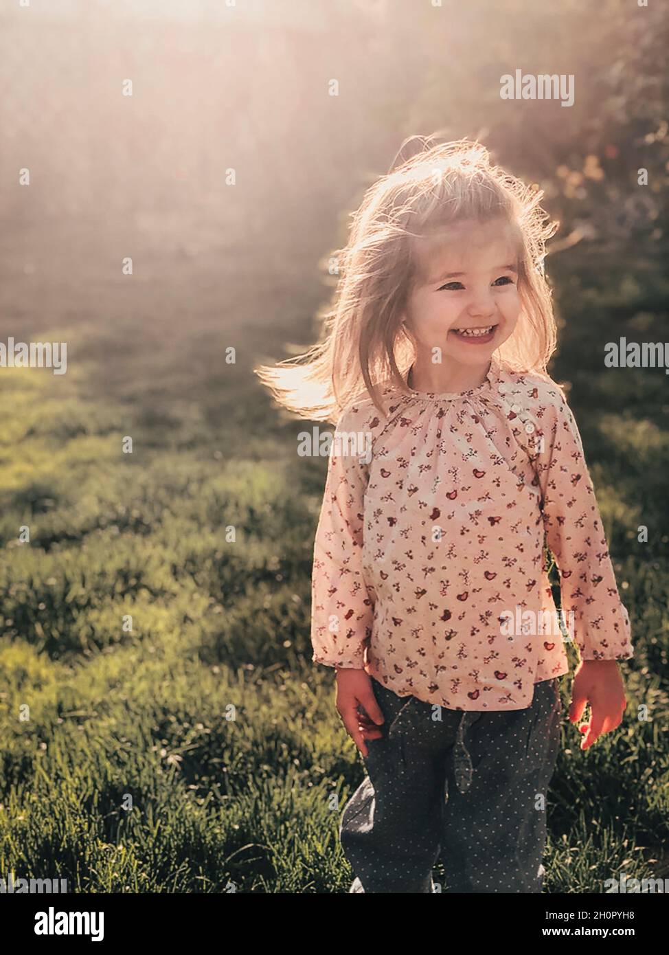 Close up photo of smiling cute little girl of 3-4 years old with blond fluffy hair stands outdoor in bright warm sunlight against backdrop of spring n Stock Photo