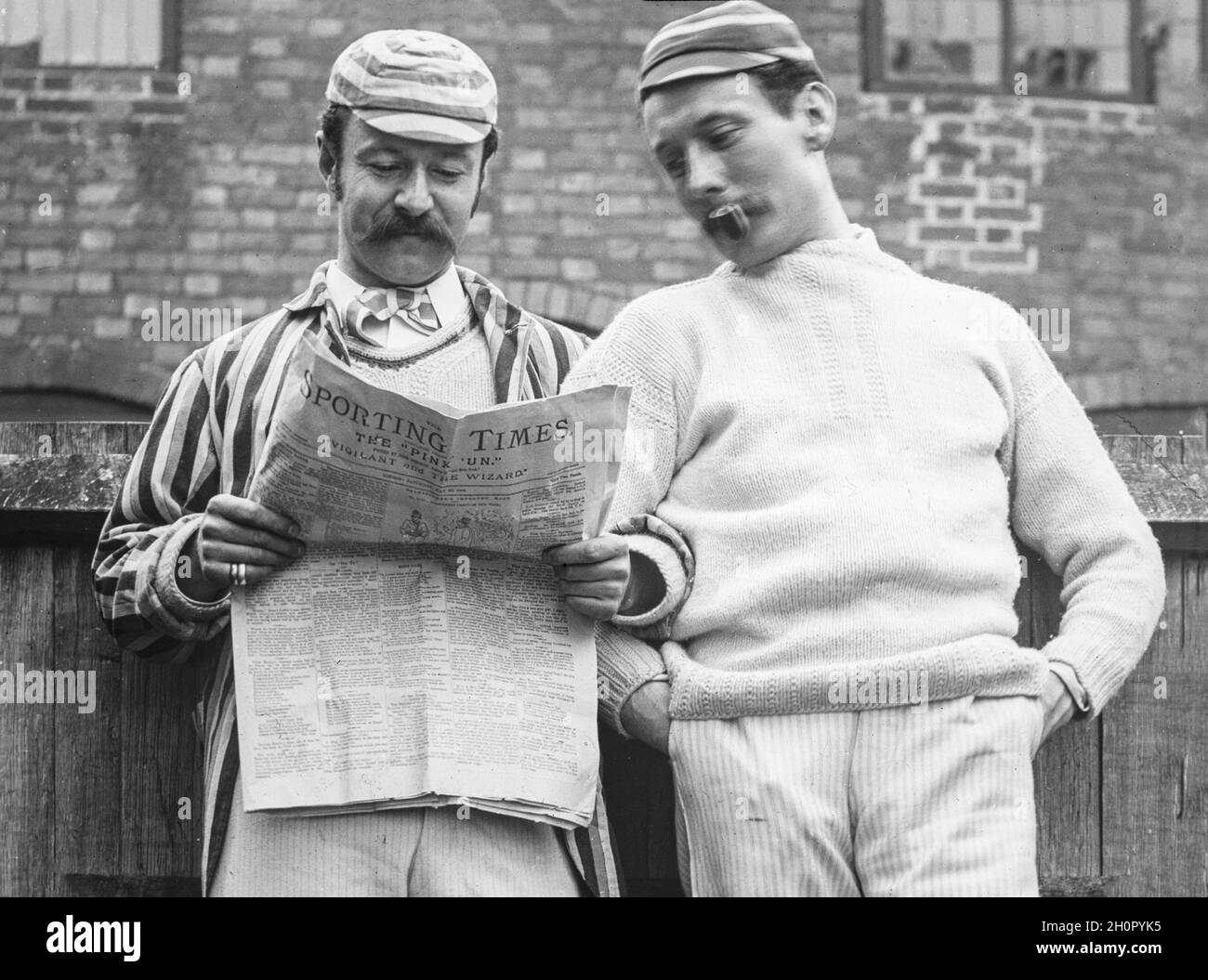 Vintage late Victorian black and white English photograph showing two men dressed in period sporting attire, maybe for cricket or rowing, reading a copy of the Sporting Times newspaper, known as the Pink 'Un. One man smoking a pipe. Stock Photo