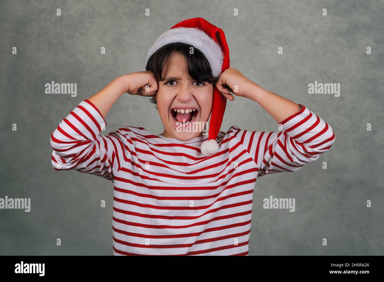 Angry kid wearing Santa Claus hat with his hands on his head over gray background Stock Photo