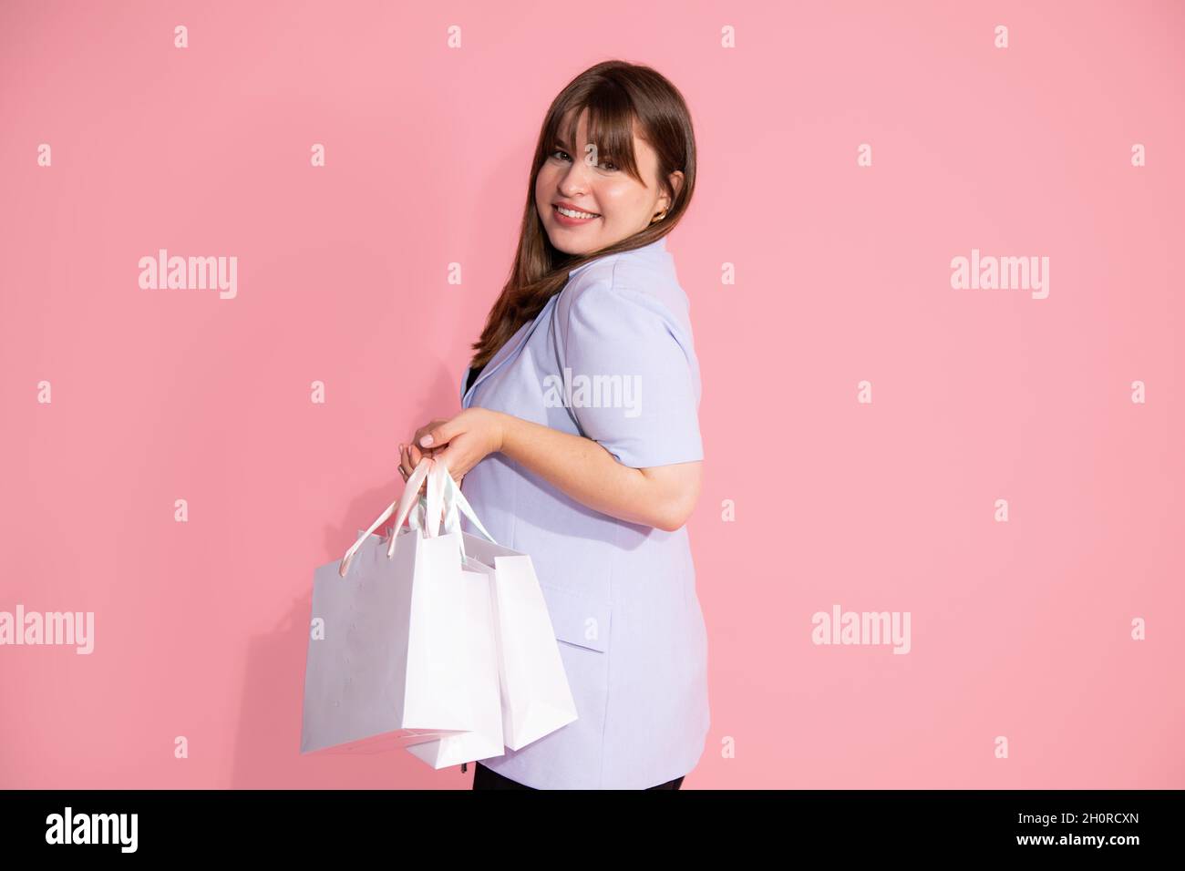 cheerful brunette young woman with bangs holdsshopping bags with packages on pink background in studio. Stock Photo
