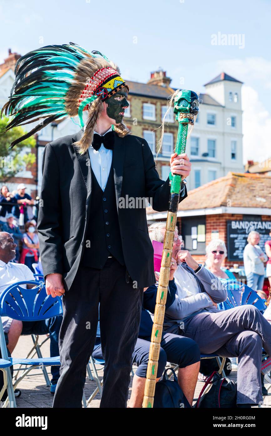 Man standing in black suit, bowtie and wearing an indian feather bonnet with audience behind. The fool from Brooms Bricks and Bowlers morris side. Stock Photo