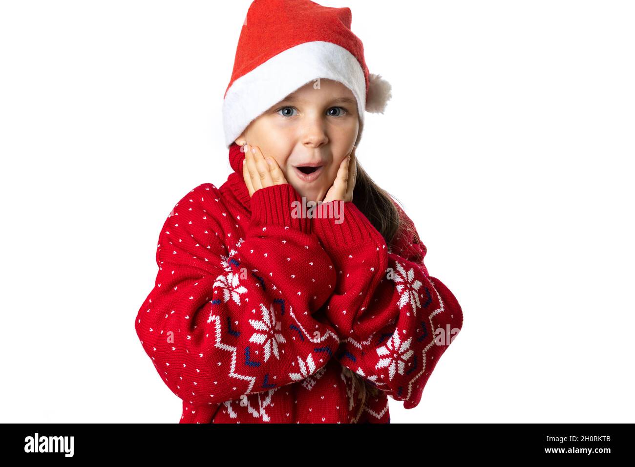 portrait of excited, charming girl in red Santa Claus hat in red Christmas sweater with reindeer touching face with palms, isolated on white Stock Photo