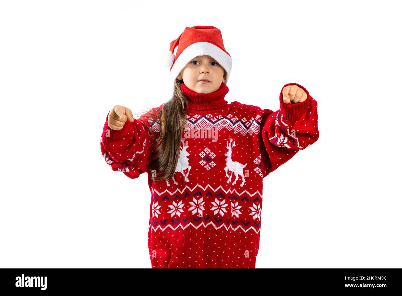 portrait of six-year-old girl in red Santa Claus hat in red knitted Christmas sweater with reindeer pointing fingers at camera, isolated on white Stock Photo