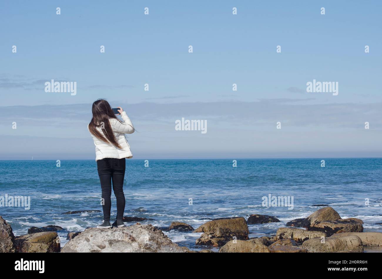 Rear view of a teenage girl standing on a rock taking a photo, Mar del Plata, Buenos Aires Province, Argentina Stock Photo